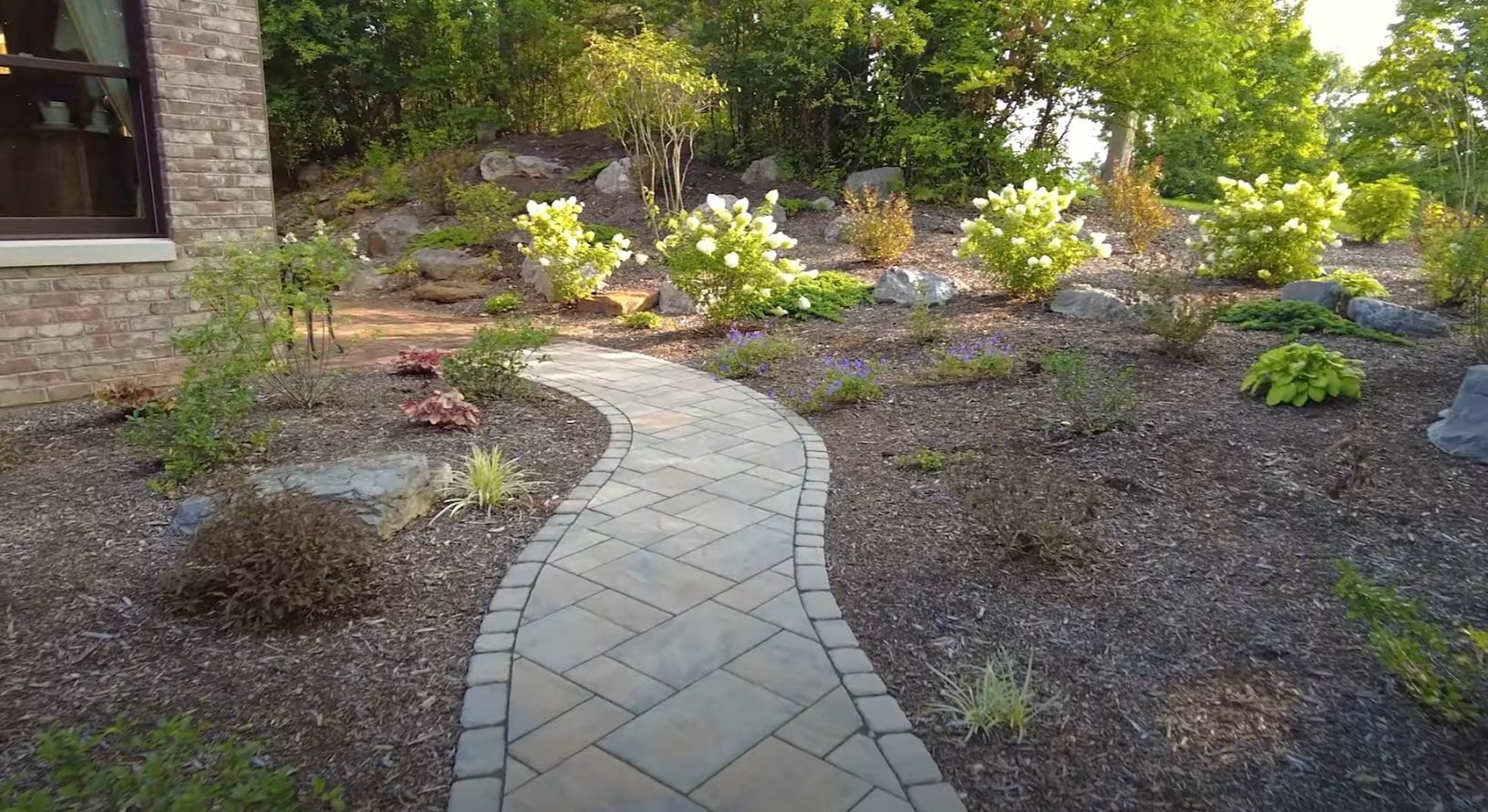 Custom hardscaping design featuring elegant curved pathways and native plant installations Curved stone pathway through professionally landscaped garden with decorative rocks and white flowering shrubs
